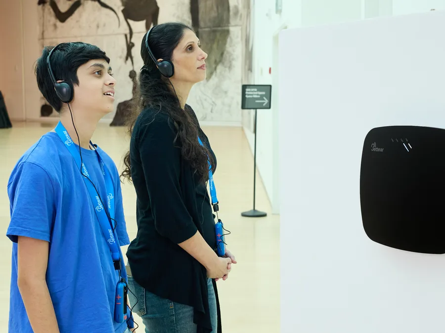 Two visitors listening on headphones beside a CASTER on the gallery wall